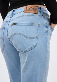 Ανοιχτό μπλε τζιν Lee με πίσω τσέπες και λογότυπο, φορεμένο με μαύρη μακρυμάνικη μπλούζα, εμφανιζόμενο από τη μέση μέχρι το μέσο του μηρού.