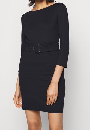 Femme portant une robe noire cintrée avec des manches trois-quarts et une large ceinture assortie avec une boucle ronde à la taille.