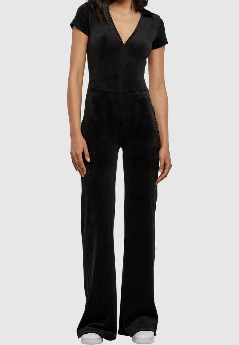 Femme portant une combinaison noire à manches courtes avec une fermeture éclair à l'avant, un pantalon à jambes larges et des baskets blanches sur un fond uni.