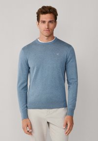 Lichtblauwe gebreide trui met een ronde hals, geribde manchetten en zoom, en een klein logo op de borst. Draagt beige broek.