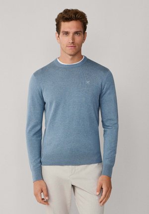 Jersey azul claro de punto con cuello redondo, puños y dobladillo acanalados, y un pequeño logo en el pecho. Se lleva con pantalones beige.