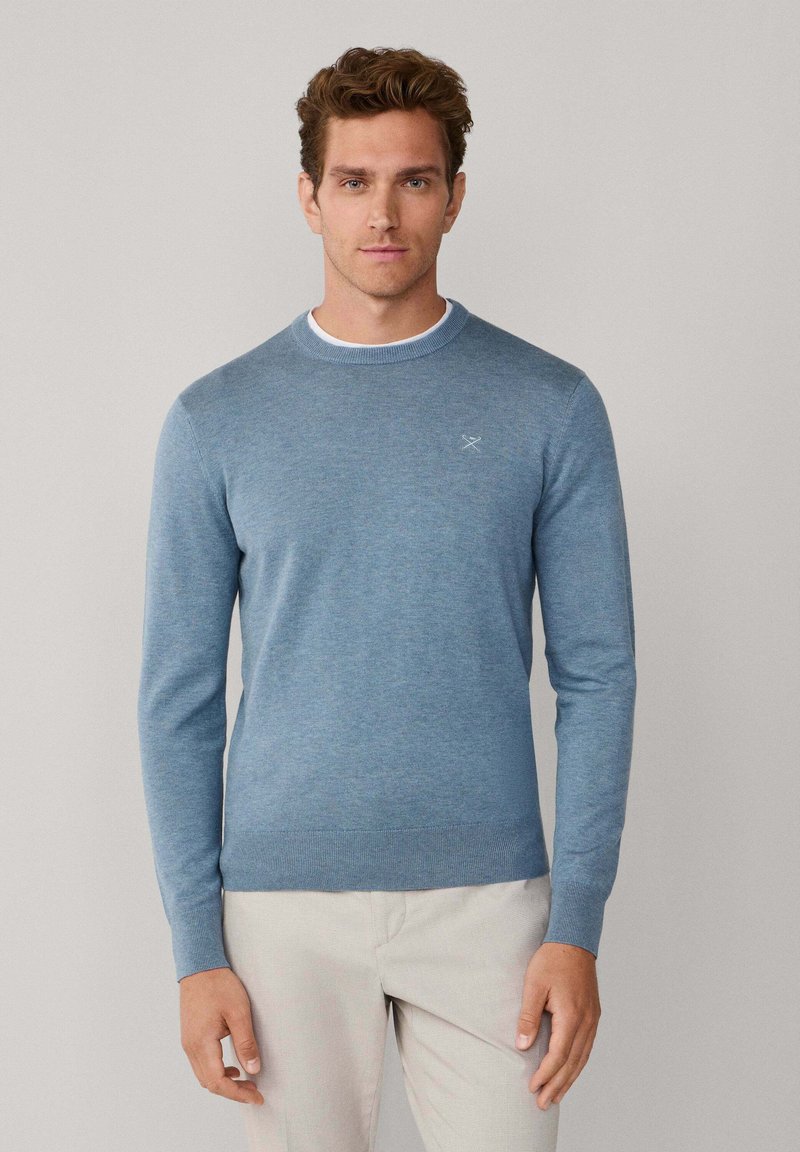 Lichtblauwe gebreide trui met een ronde hals, geribde manchetten en zoom, en een klein logo op de borst. Draagt beige broek.