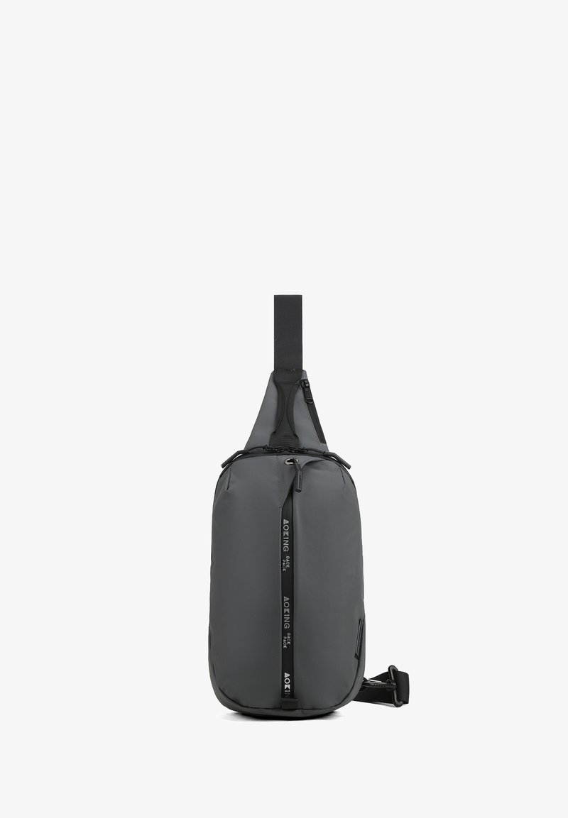 Sac à dos bandoulière noir avec une poche zippée avant, une sangle ajustable et une tirette de fermeture éclair verticale marquée.