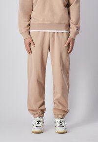 Pantalones de chándal beige claro con puños elásticos y corte relajado. Llevados con zapatillas blancas y negras con acentos distintivos.
