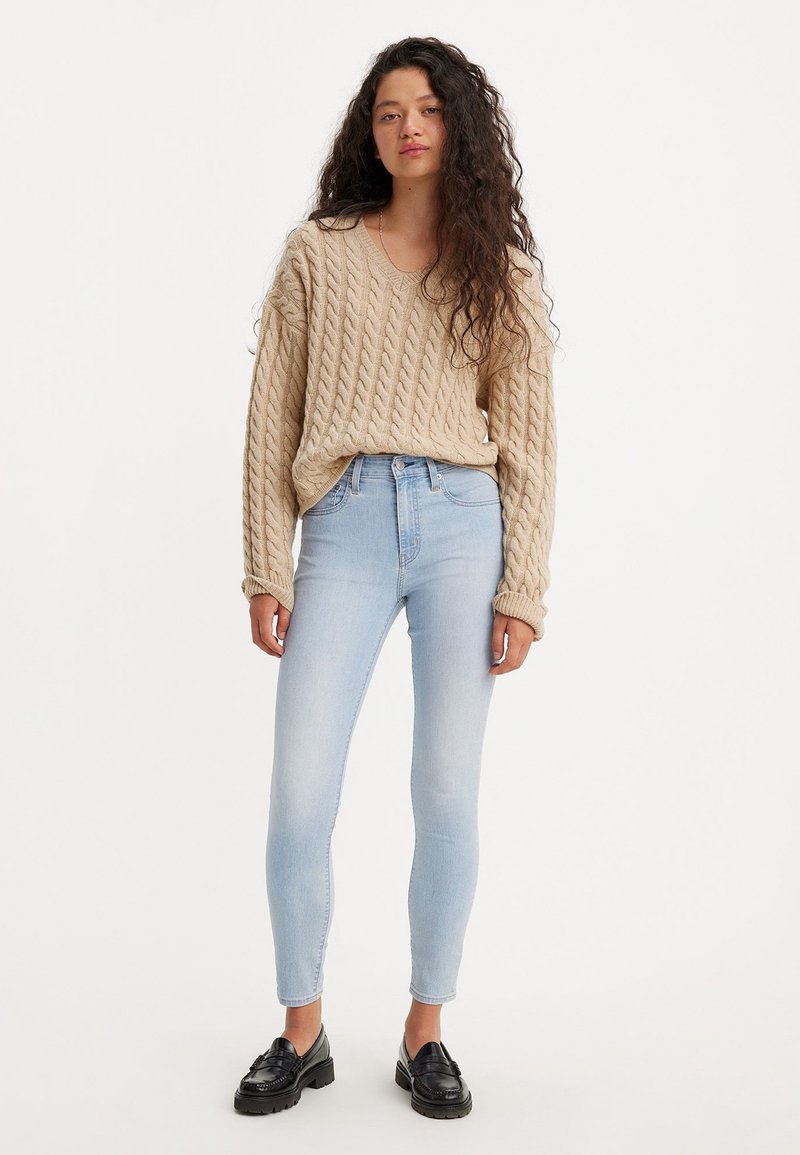 Pull en tricot beige avec un col en V, associé à un jean skinny bleu clair et des mocassins noirs. Texture lisse et coupe décontractée.