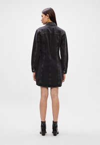 Vestido de ganga preto com mangas longas, colarinho e detalhes em botões. Apresenta um corte reto com costuras decorativas e comprimento até a metade das coxas.