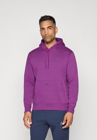 Lilla hoodie med en frontlomme og snørehætte. lavet af blødt stof, med et lille sort logo på brystet. Parret med mørkeblå bukser.