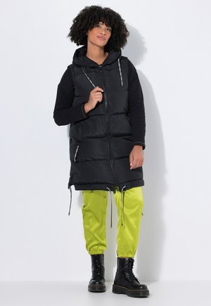 Chaleco tipo pluma negro con capucha, que cuenta con bolsillos con cremallera y cordones. Combinado con pantalones ajustados de un verde brillante y botas negras con cordones.