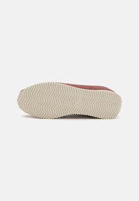 Semelle de chaussure avec motif de bande de roulement zigzag beige vue de dessous, partie de la tige rouge visible sur fond blanc.