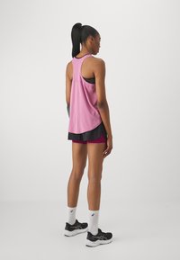 Tumehobusega naine, kes kannab roosa ilma varrukateta topsi, musta spordibrassi, kihilisi musti ja magenta lühikesi pükse, valgeid sokke ja musti jooksujalatseid, seisab.