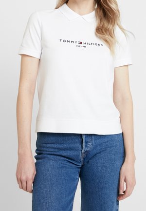 Vrouw in een witte Tommy Hilfiger polo met logo en blauwe spijkerbroek, getoond van schouders tot heupen tegen een effen achtergrond.