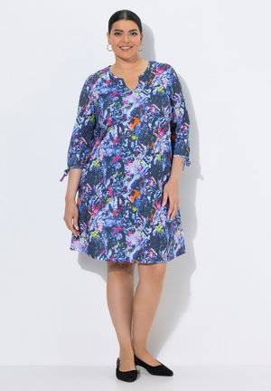 Femme portant une robe bleue à imprimé abstrait longueur genou avec manches 3/4 et des chaussures plates noires, debout sur un fond blanc.