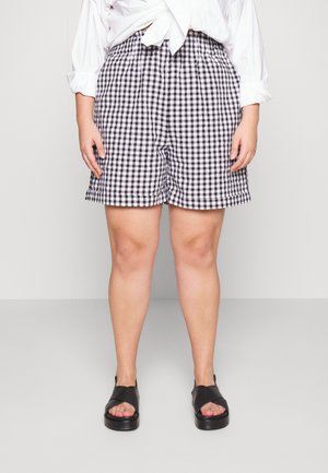 Kvinde står iført sort-hvid gingham-shorts, hvid langærmet skjorte bundet foran og sorte åbne sandaler på en grå gulv.