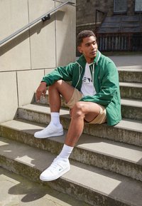 Zelená větrovka, bílé tričko s logem, béžové šortky, bílé tenisky s logem, sedící na šedých schodech s odrazy stínů od zábradlí.