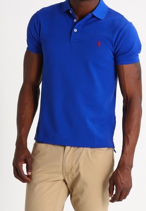 Niebieska koszulka polo z bawełny z kołnierzykiem, krótkimi rękawami i małym czerwonym logo na piersi. Zestawiona z beżowymi spodniami.