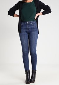 Frau steht mit den Händen in den Hüften, trägt dunkelblaue Skinny-Jeans, schwarze Stiefeletten, grünes Top und schwarzen Cardigan vor weißem Hintergrund.