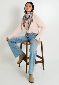Un pull rose clair avec un tricot texturé, un jean bleu évasé, des bottines beiges et une écharpe à motifs avec des tons de bleu et de violet, assis sur un tabouret.