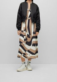 Svart bomberjacka över en mönstrad, beige och vit midiklänning med mörka linjer. Matchas med vita sneakers och mönstrade strumpor.