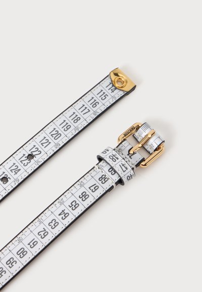 Ceinture conçue pour ressembler à un ruban à mesurer blanc avec des chiffres noirs et une boucle dorée sur un fond clair.
