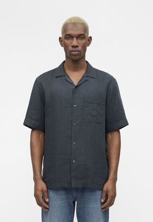 Muž s krátkymi blond vlasmi, ktorý má na sebe čiernu krátkym rukávom zapínanú košeľu a modré džínsy, stojaci pred jednoduchým bielym pozadím.