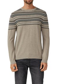 Man in een beige gebreid sweater met zwart, wit en groene geometrische patronen op de borst en schouders, gecombineerd met donkere jeans.