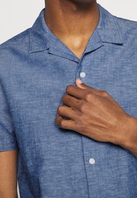 Camicia azzurra a maniche corte con bottoni, realizzata in tessuto testurizzato, con colletto e bottoni bianchi, indossata da una persona che tiene il colletto.