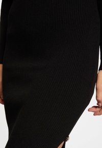 Abito maglione nero a coste con maniche lunghe, caratterizzato da uno spacco laterale e un dettaglio in tono oro sull'orlo. Tessuto a maglia morbido, design aderente.