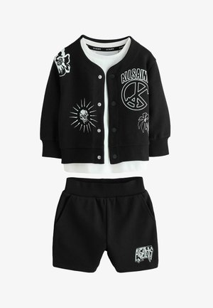 Czarna kurtka zapinana na guziki z nadrukami białych czaszek, znaku pokoju i kwiatów, w zestawieniu z białą koszulą pod spodem oraz czarnymi szortami z nadrukiem logo.