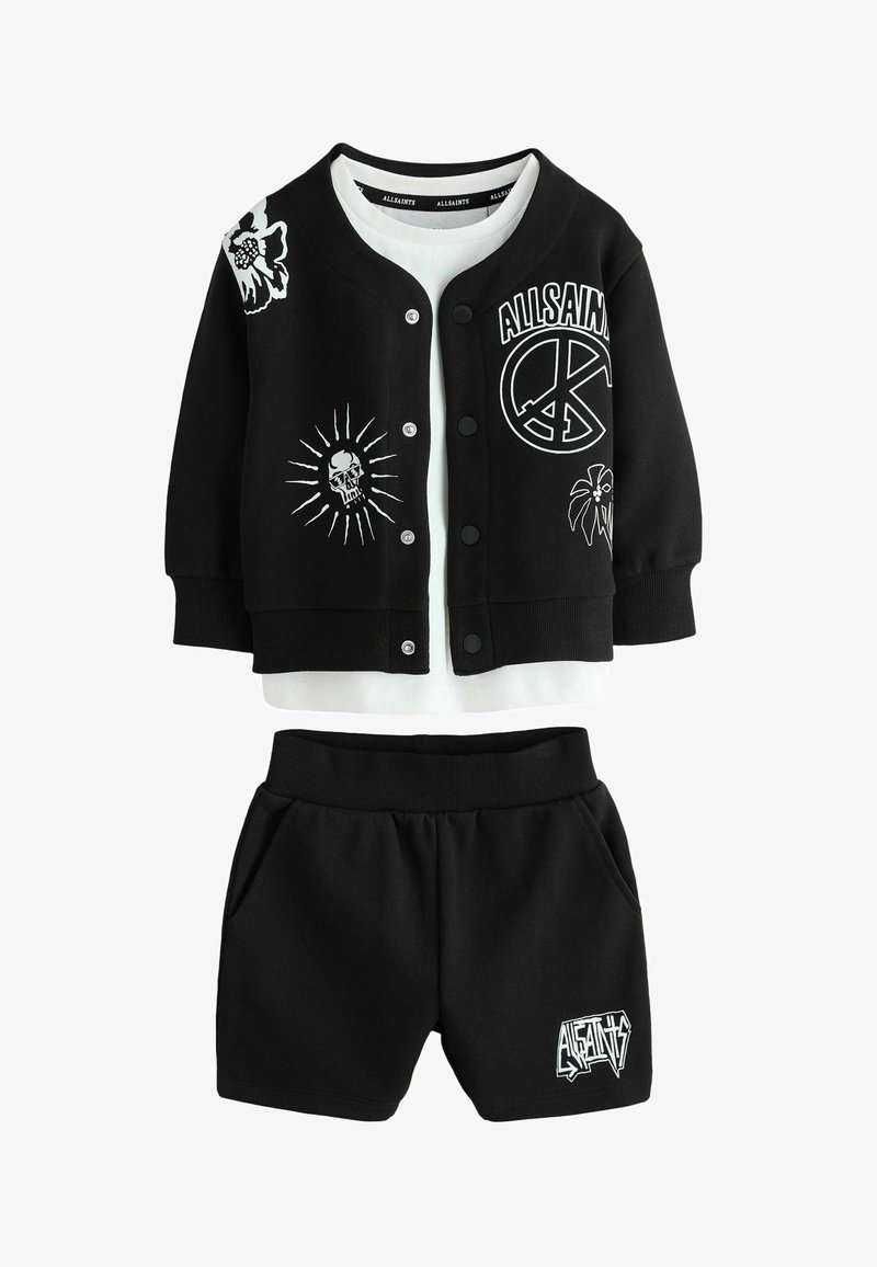 Chaqueta negra con botones y estampados en blanco de calaveras, símbolo de la paz y flores, combinada con una camiseta blanca debajo y pantalones cortos negros con estampado de logotipo.