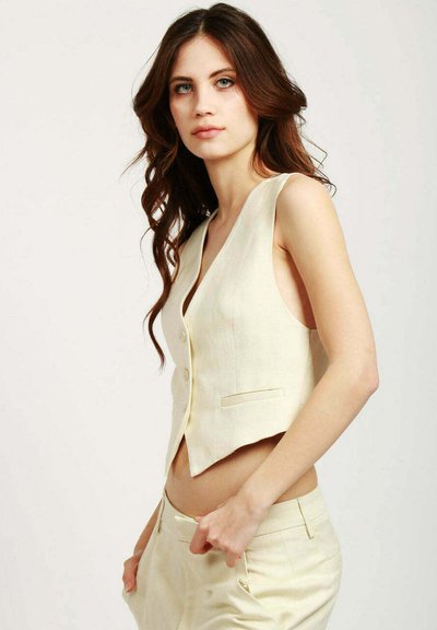 Gilet in lino color crema con scollatura a V profonda, due bottoni e tasche laterali, abbinato a pantaloni chiari coordinati.