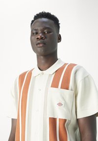 Camisa polo de manga corta para hombre en crema con rayas verticales en naranja y beige, que cuenta con un cuello abotonado y un bolsillo en el pecho con el logo.