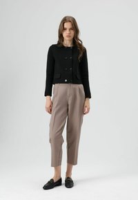 Zwarte cropped jas met ronde hals en knoopsluiting, gecombineerd met beige geplooide broek en zwarte loafers. Overal gladde texturen.