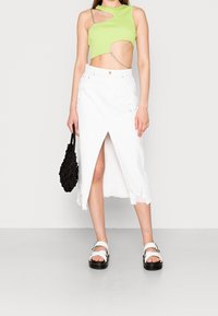 Top court vert avec un design asymétrique et un détail de chaîne, associé à une jupe en denim blanche avec une fente à l'avant, un ourlet effiloché et un sac noir.