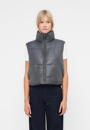 Grå puffervest med høy krage, glidelåslukking og horisontal søm. Bært over en hvit skjorte, kombinert med mørke denimbukser.