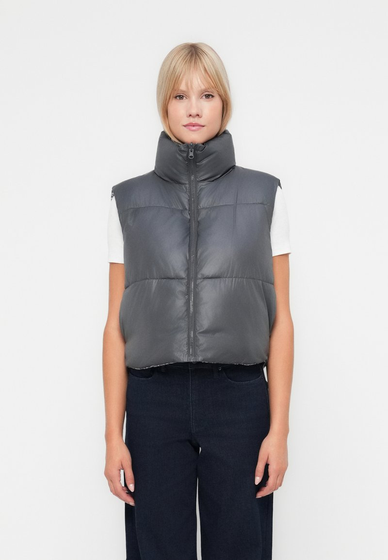 Colete puffer cinza com colarinho alto, fecho de correr e costura horizontal. Usado sobre uma camisa branca, combinado com calças de ganga escuras.