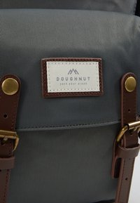 Sac à dos en tissu gris avec une accent en sangle en cuir, quincaillerie dorée et un patch cousu étiqueté "DOUGHNUT" avec des graphiques de montagne.