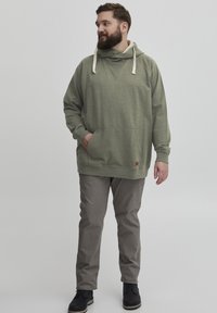 Sudadera verde oliva con capucha ajustable, bolsillo frontal y puños acanalados, combinada con pantalones gris claro y botas negras. Diseño minimalista.