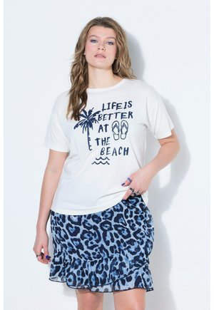 Mujer con camiseta blanca con texto "La vida es mejor en la playa" y gráfico de palmera, combinada con falda azul con estampado de leopardo.