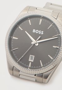 Ανοξείδωτο ατσάλινο ρολόι χειρός με μαύρο καντράν, ασημένιους δείκτες ώρας, παράθυρο ημερομηνίας στις 6 η ώρα και το λογότυπο "BOSS" κάτω από τις 12 η ώρα.