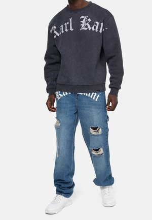 Felpa grigio scuro con grande testo gotico bianco e jeans blu strappati con scarpe da ginnastica bianche indossati da una persona in piedi su sfondo bianco.