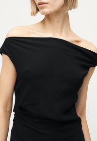 Femme portant un haut noir épaules dénudées, montrant les clavicules et une partie des bras supérieurs sur un fond clair uni.