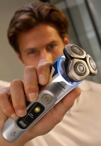 Philips scheerapparaat met een zilveren behuizing, drie ronde metalen koppen, blauwe LED-lamp en bedieningsknoppen voor voeding en instellingen.