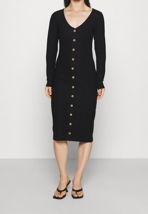 Černé žebrované šaty s dlouhými rukávy a hlubokým výstřihem do V, s řadou zlatých knoflíků po straně přední části. Kombinovány s černými žabkami.