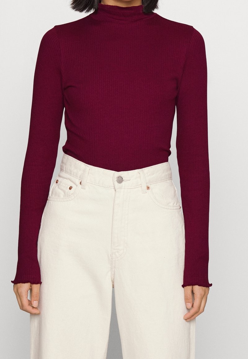 Femme portant un pull à col roulé côtelé bordeaux ajusté, rentré dans un jean écru taille haute avec poches avant et fermeture par boutons.