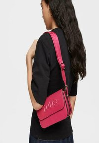 Bolso bandolera rosa con superficie texturizada, logotipo "TOUS" en relieve, correa ajustable y cierre de cremallera, llevado en el hombro.