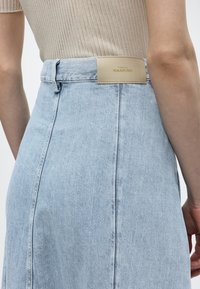 Κοντινό πλάνο ενός ατόμου που φοράει φούστα από ανοιχτό μπλε τζην με μπεζ ετικέτα "7 For All Mankind" στη μέση και μπεζ ριγέ τοπ.