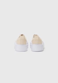 Beige vászon slip-on sportcipők vastag fehér gumitalppal és saroklehúzóval. Az outsole oldalán domború logó található.