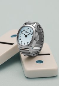 Ρολόι χειρός Timex με ασημένιο μεταλλικό λουράκι δείχνει 10:10, ακουμπισμένο σε δύο λευκά κεραμικά ντόμινο με μαύρες τελείες σε ανοιχτό γαλάζιο φόντο.