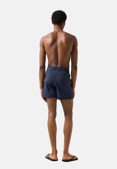 Mand stående barfodet med ryggen til, iført marineblå badeshorts og sorte flip-flops mod en ensartet lys baggrund.