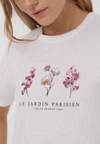 Maglietta bianca in cotone con stampa floreale nei toni del rosa e del viola, con la scritta "LE JARDIN PARISIEN" e un design grafico.